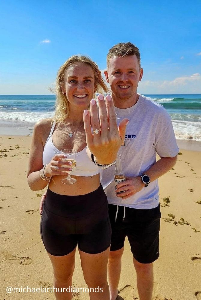 cheap proposal ideas happy couple standing on sandy beach with woman showing oval engagement ringІ michaelarthurdiamonds