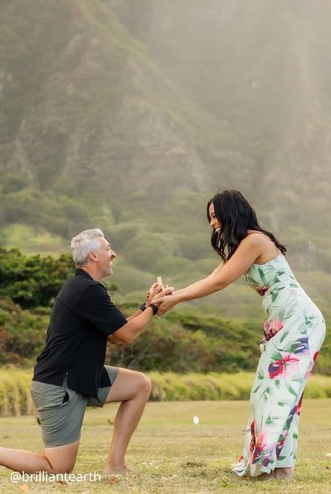 cheap proposal ideas man proposing on one knee to smiling woman in floral dress in green valley brilliantearth