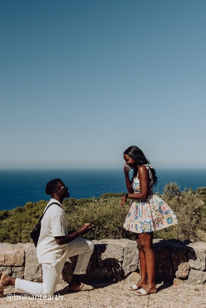 cheap proposal ideas man proposing on one knee to surprised woman by stone wall overlooking blue ocean brilliantearth