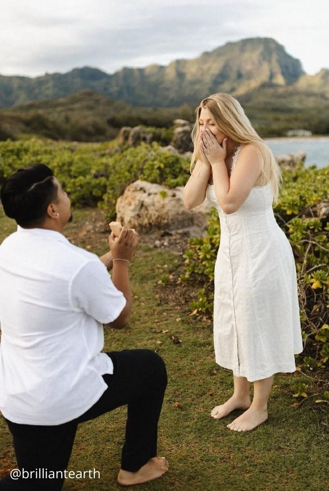 cheap proposal ideas man proposing on one knee to surprised woman in white dress on coastal cliff brilliantearth