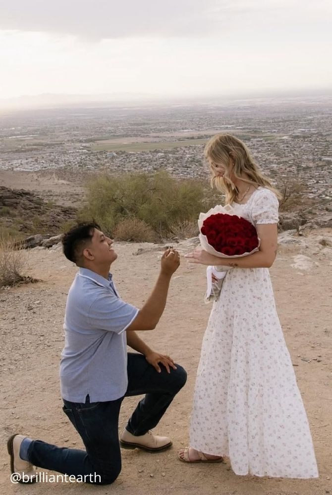 cheap proposal ideas man proposing on one knee to woman holding red roses on scenic hilltop brilliantearth