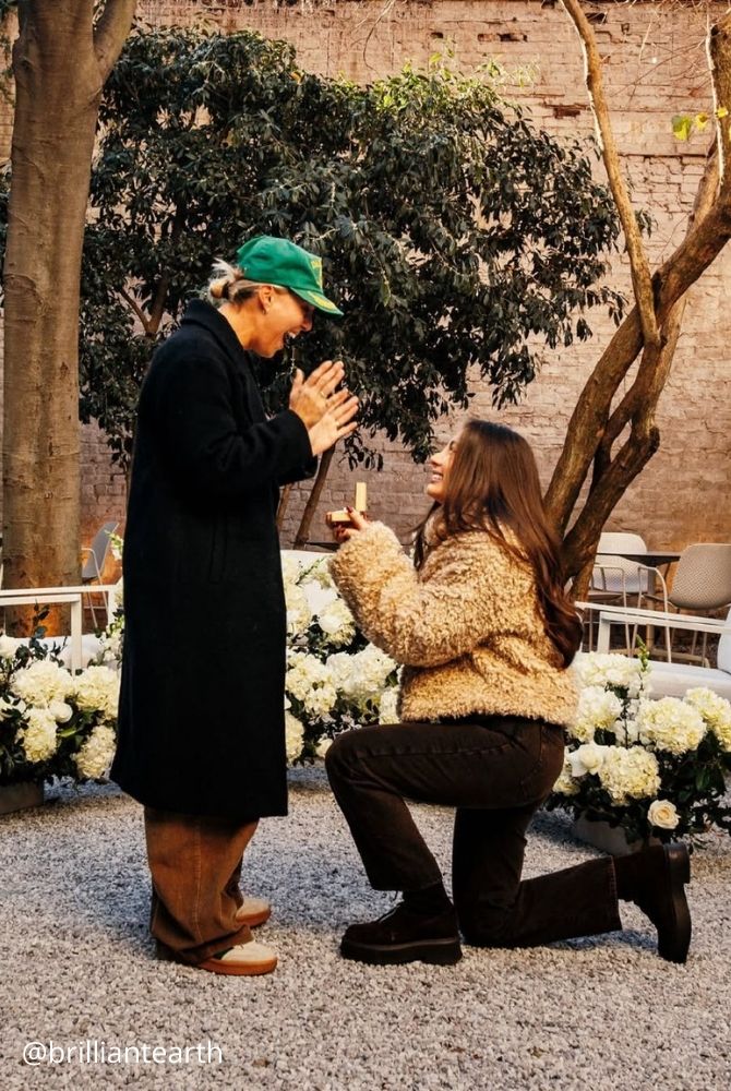 cheap proposal ideas woman proposing on one knee to surprised partner in courtyard with white flowers brilliantearth